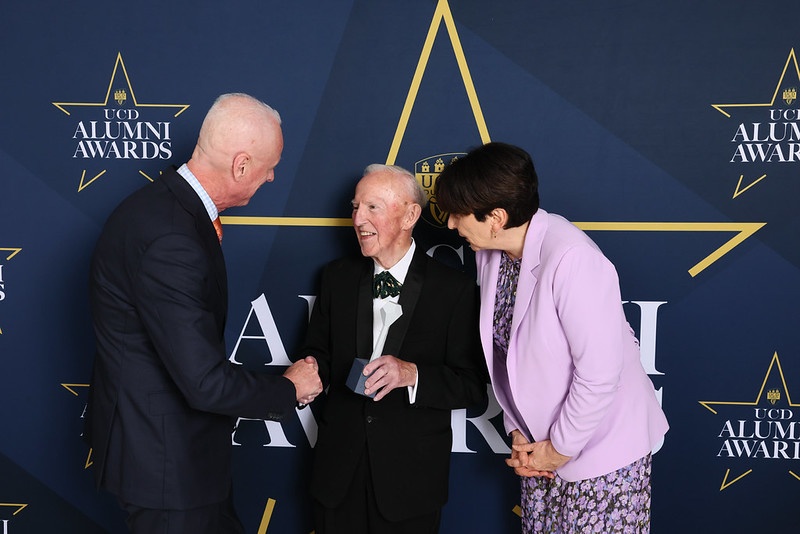 Three people stand in front of a UCD Alumni Awards backdrop, Peter Timoney in a tuxedo shaking hands with another man while President Orla Feely in a lilac blazer smiles beside them.